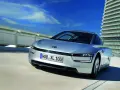 Srebrny, futurystyczny VW XL1 sunie po drodze obok nowoczesnego budynku.