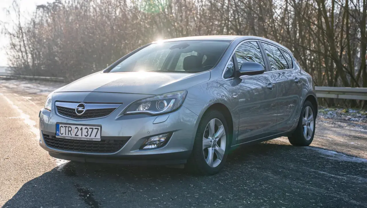 Szary Opel Astra J hatchback na tle drzew i słońca.