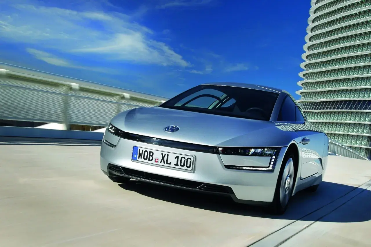 Srebrny, futurystyczny VW XL1 sunie po drodze obok nowoczesnego budynku.