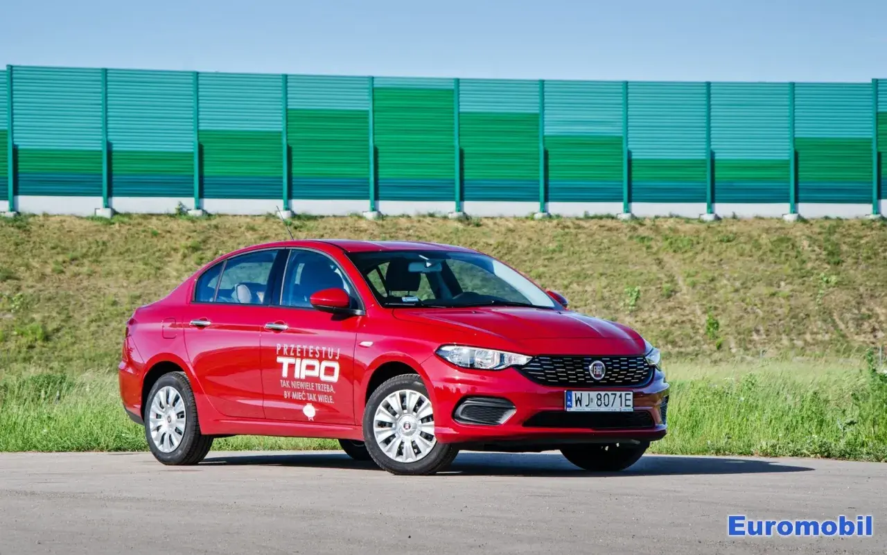 Czerwony fiat tipo sedan z napisem "Przetestuj Tipo" na drzwiach, zaparkowany na tle zielonego ekranu dźwiękochłonnego.