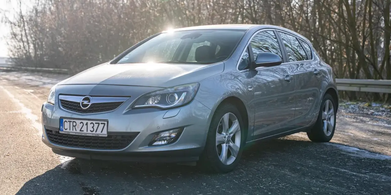 Szary Opel Astra J hatchback na tle drzew i słońca.