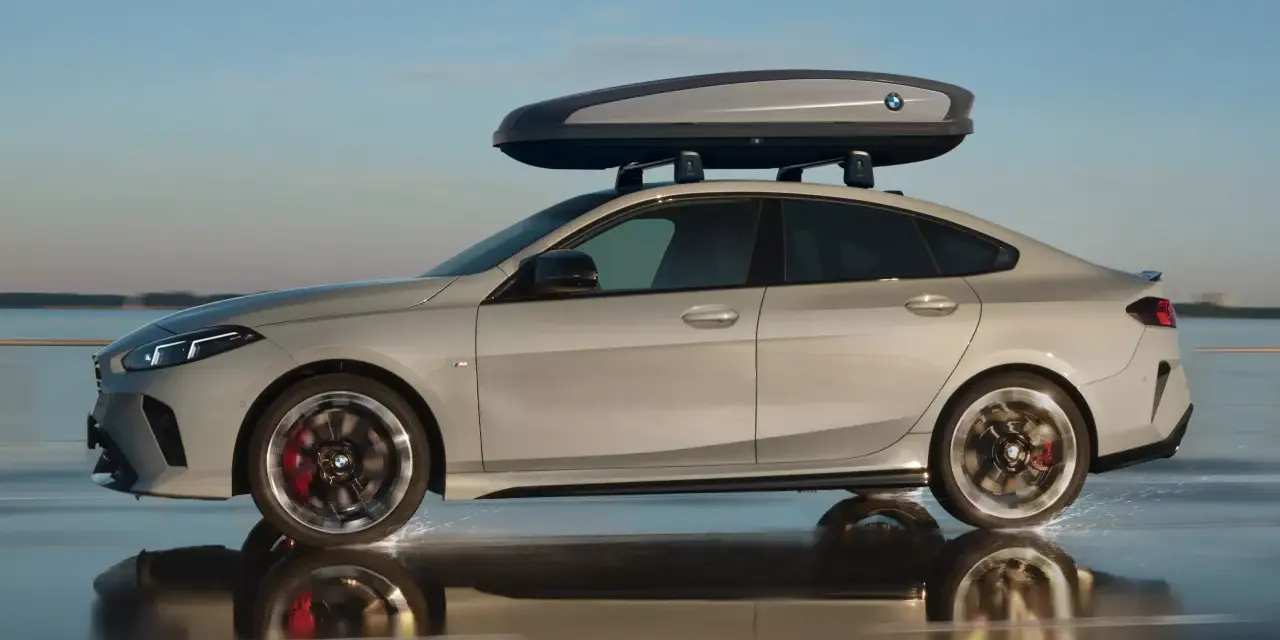 Srebrne BMW Gran Coupé z boxem dachowym na relingach jedzie po mokrej nawierzchni, rozpryskując wodę.