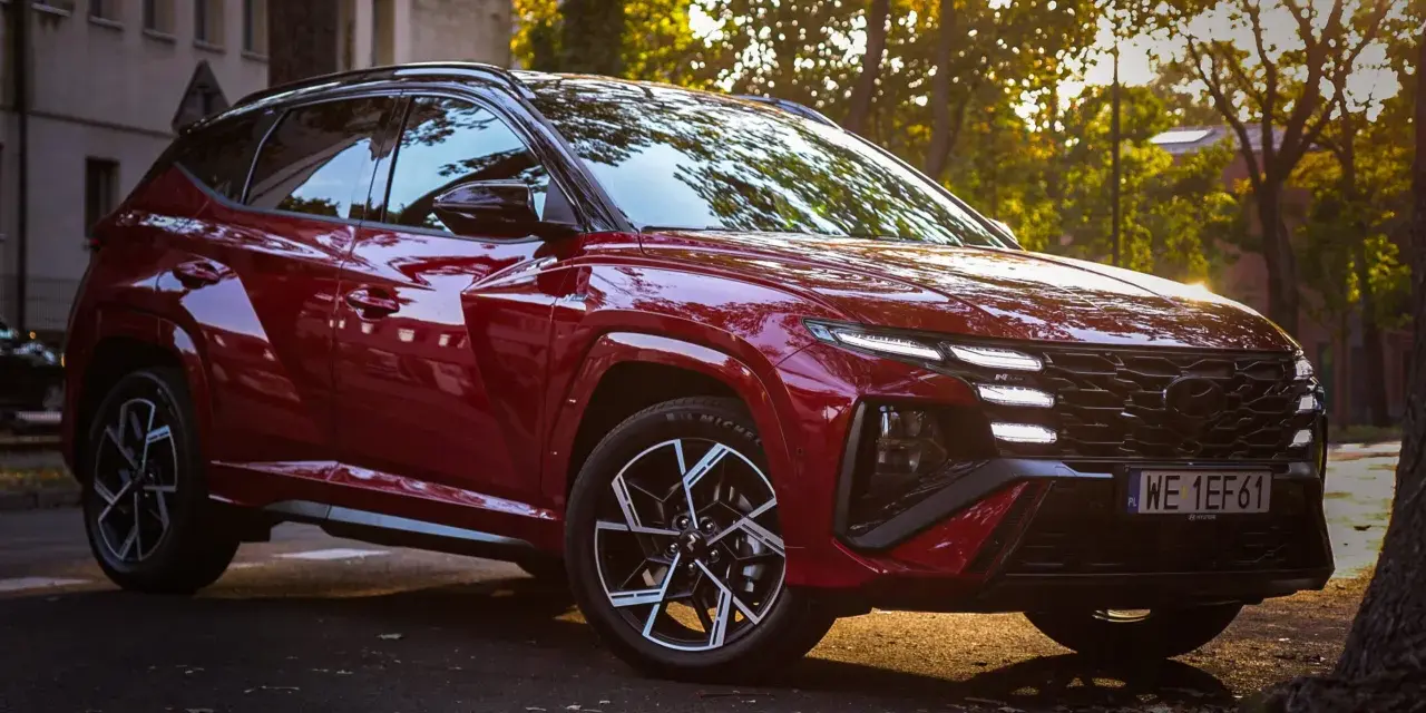 Czerwony Hyundai Tucson hybryda zaparkowany na tle jesiennych drzew i kamienicy.
