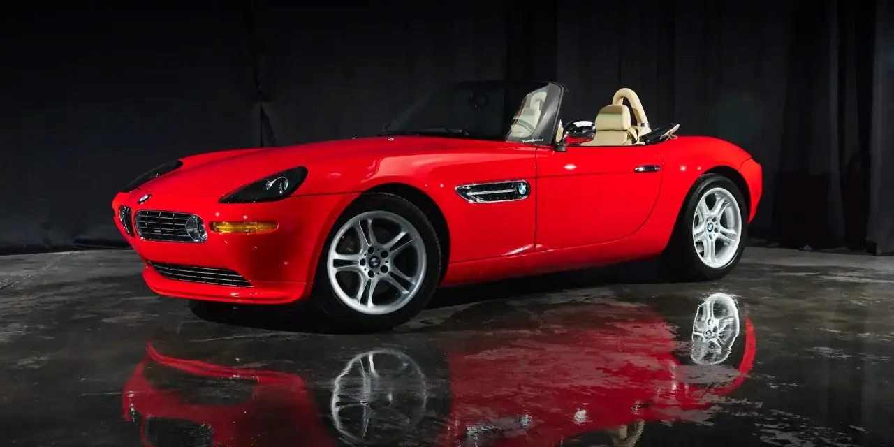 Czerwony kabriolet BMW Z8 z beżowym wnętrzem, lśniący na mokrej podłodze, odbija się w tafli wody.