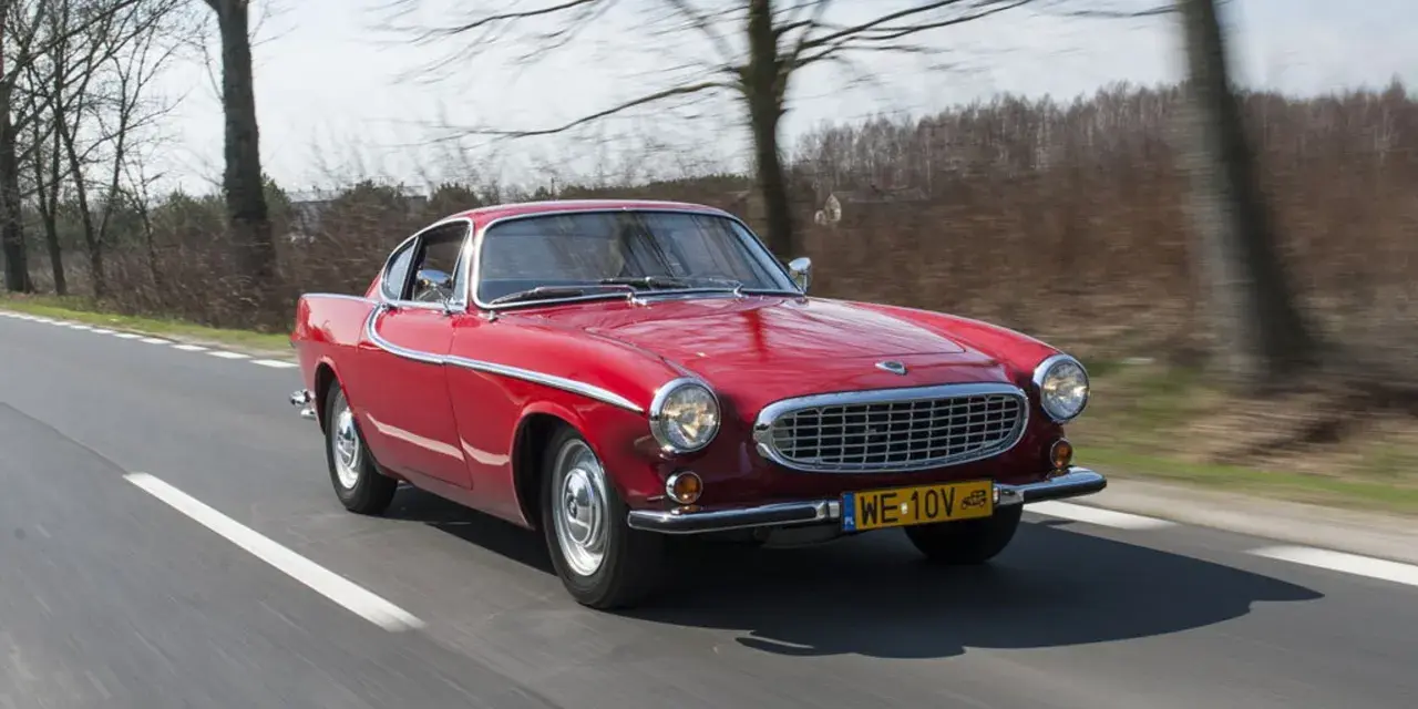 Czerwone Volvo P1800 sunie po drodze, otoczone drzewami. Klasyk z lat 60. w pełnej krasie.