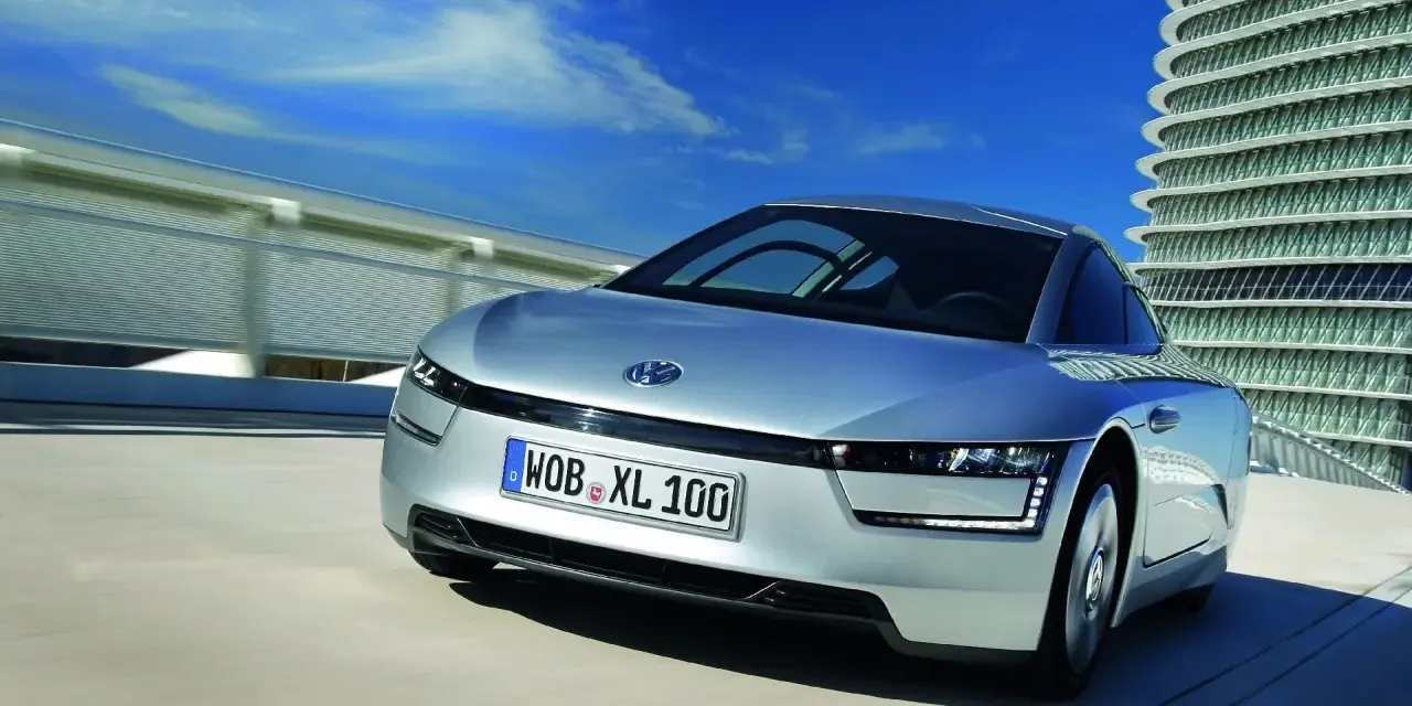 Srebrny, futurystyczny VW XL1 sunie po drodze obok nowoczesnego budynku.
