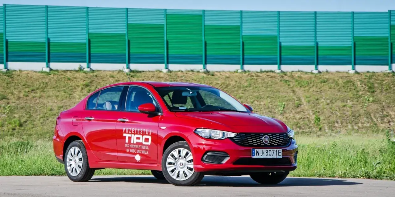 Czerwony fiat tipo sedan z napisem "Przetestuj Tipo" na drzwiach, zaparkowany na tle zielonego ekranu dźwiękochłonnego.