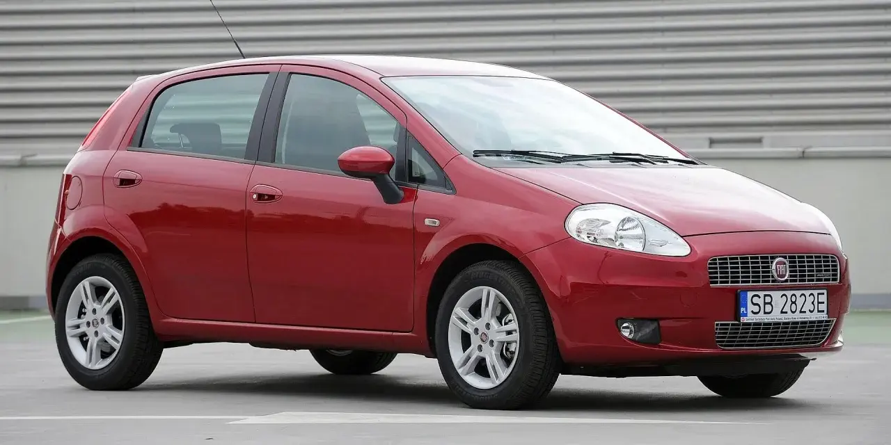 Czerwony Fiat Punto Evo z polskimi tablicami SB 2823E zaparkowany na szarym podłożu, z metalową ścianą w tle.