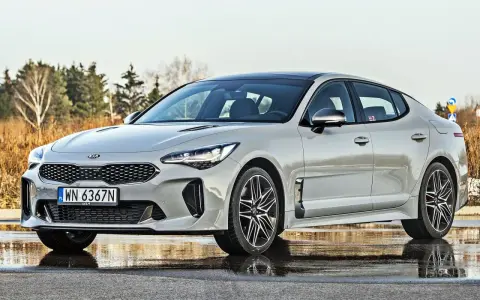 Elegancka Kia Stinger 2025 w kolorze srebrnym, zaparkowana na mokrej nawierzchni, z drzewami w tle.