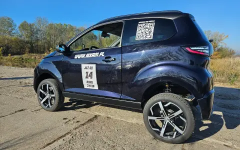 Ciemnoniebieskie Aixam auto z naklejkami "JUŻ OD 14 ROKU ŻYCIA" i kodem QR stoi na tle jesiennego krajobrazu.