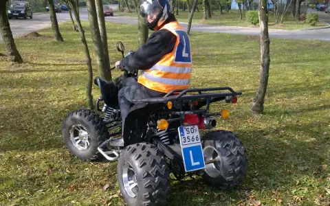 Młody kierowca w kamizelce odblaskowej i kasku uczy się jeździć quadem. Tablica z literą "L" wskazuje, że to kategoria B1.