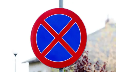 Ostrzeżenie dla kierowców: zakaz parkowania. Znak drogowy w kształcie koła z czerwonym obramowaniem i niebieskim tłem, na którym widnieje czerwony krzyż.