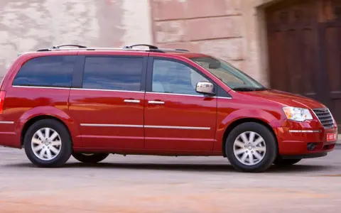 Czerwony Chrysler Grand Voyager w ruchu, z przyciemnianymi szybami i relingami dachowymi, przemierza ulicę obok kamiennej fasady.