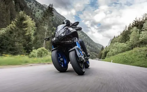 Motocyklista na Yamaha Niken pokonuje zakręt na górskiej drodze, otoczony zielenią i chmurami.