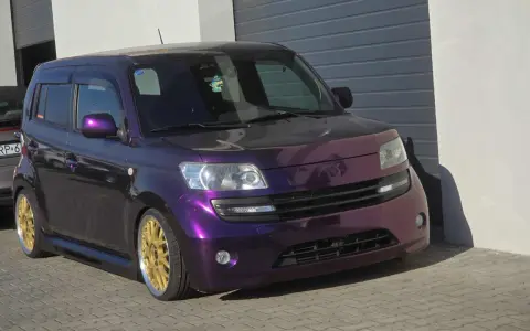 Fioletowy Daihatsu Materia z błyszczącymi złotymi felgami, zaparkowany przed garażem.