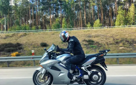 Motocyklista w kasku jedzie na srebrnym Honda VFR 800 wzdłuż leśnej drogi.