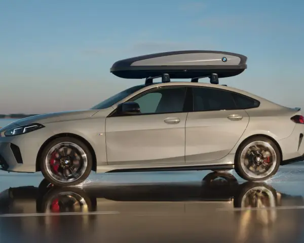 Srebrne BMW Gran Coupé z boxem dachowym na relingach jedzie po mokrej nawierzchni, rozpryskując wodę.