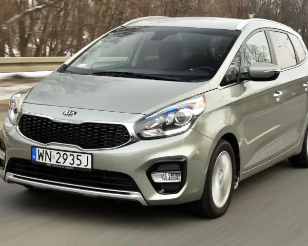 Srebrny Kia Carens, typowy MPV, jedzie po drodze. To auto rodzinne, idealne na wycieczki.