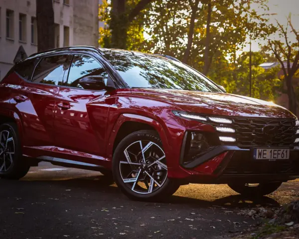 Czerwony Hyundai Tucson hybryda zaparkowany na tle jesiennych drzew i kamienicy.