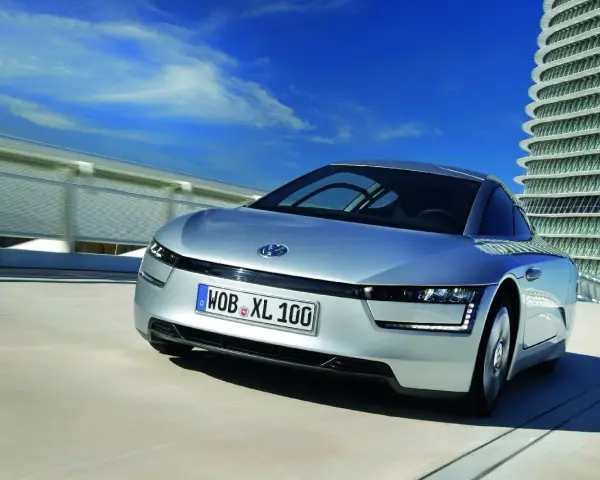 Srebrny, futurystyczny VW XL1 sunie po drodze obok nowoczesnego budynku.