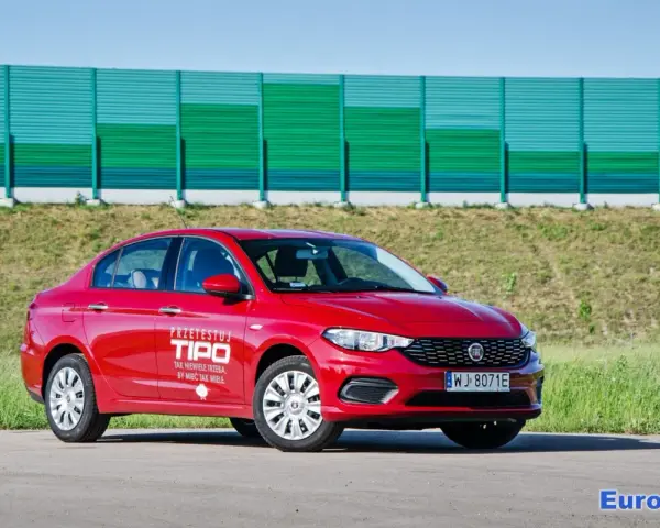 Czerwony fiat tipo sedan z napisem "Przetestuj Tipo" na drzwiach, zaparkowany na tle zielonego ekranu dźwiękochłonnego.