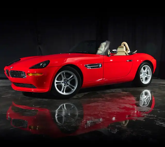 Czerwony kabriolet BMW Z8 z beżowym wnętrzem, lśniący na mokrej podłodze, odbija się w tafli wody.