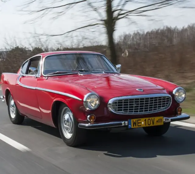 Czerwone Volvo P1800 sunie po drodze, otoczone drzewami. Klasyk z lat 60. w pełnej krasie.