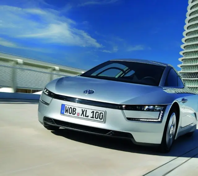 Srebrny, futurystyczny VW XL1 sunie po drodze obok nowoczesnego budynku.
