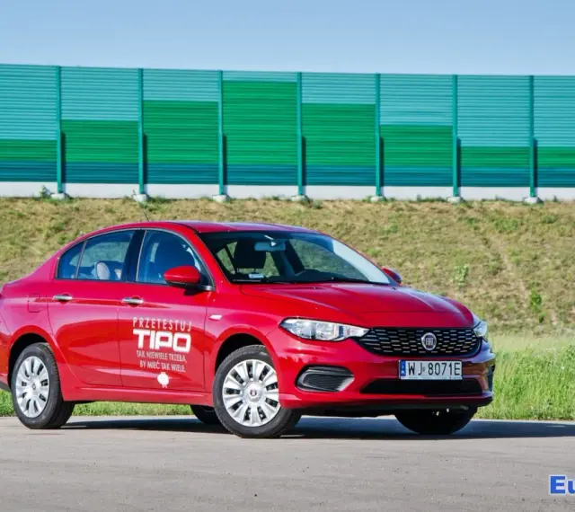 Czerwony fiat tipo sedan z napisem "Przetestuj Tipo" na drzwiach, zaparkowany na tle zielonego ekranu dźwiękochłonnego.