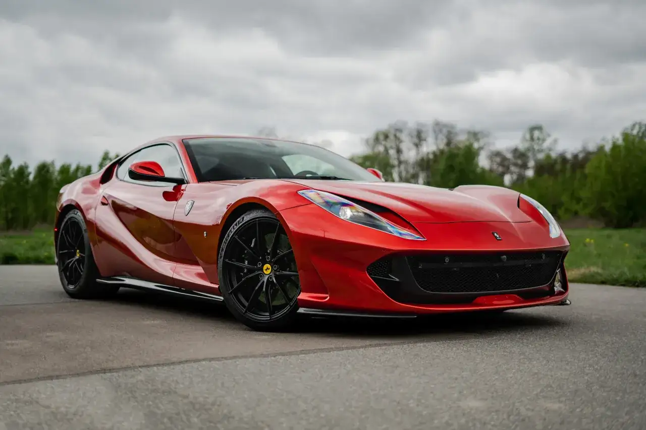 Czerwone Ferrari 812 Superfast z czarnymi felgami na tle pochmurnego nieba i zieleni.