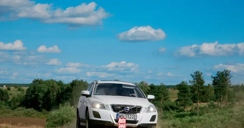Białe Volvo XC60 T6 jedzie po piaszczystej drodze wśród zieleni, pod błękitnym niebem z chmurami.