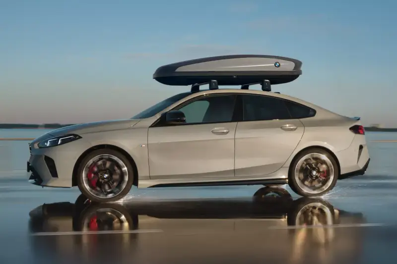 Srebrne BMW Gran Coupé z boxem dachowym na relingach jedzie po mokrej nawierzchni, rozpryskując wodę.