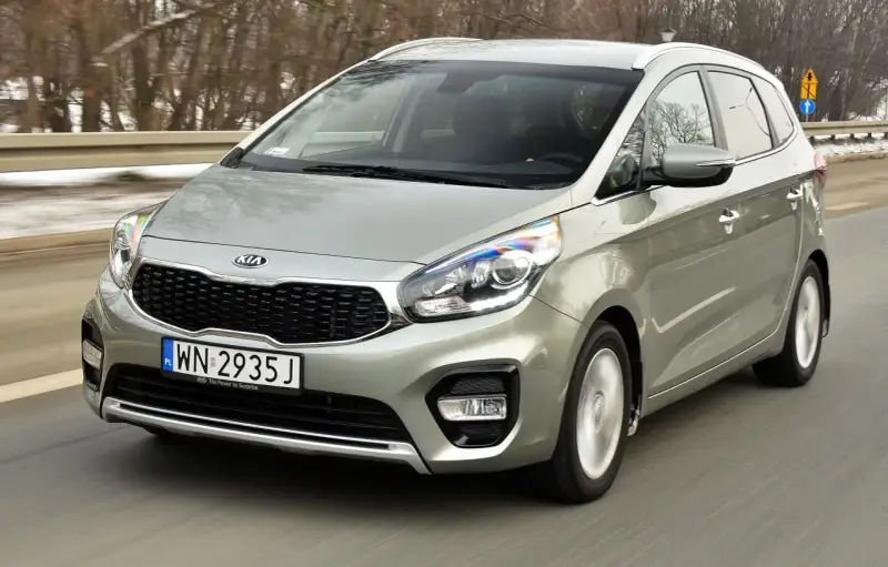Srebrny Kia Carens, typowy MPV, jedzie po drodze. To auto rodzinne, idealne na wycieczki.