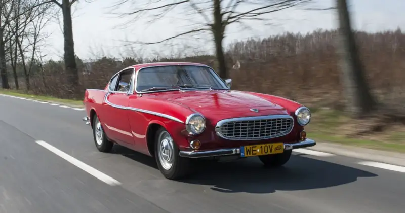Czerwone Volvo P1800 sunie po drodze, otoczone drzewami. Klasyk z lat 60. w pełnej krasie.