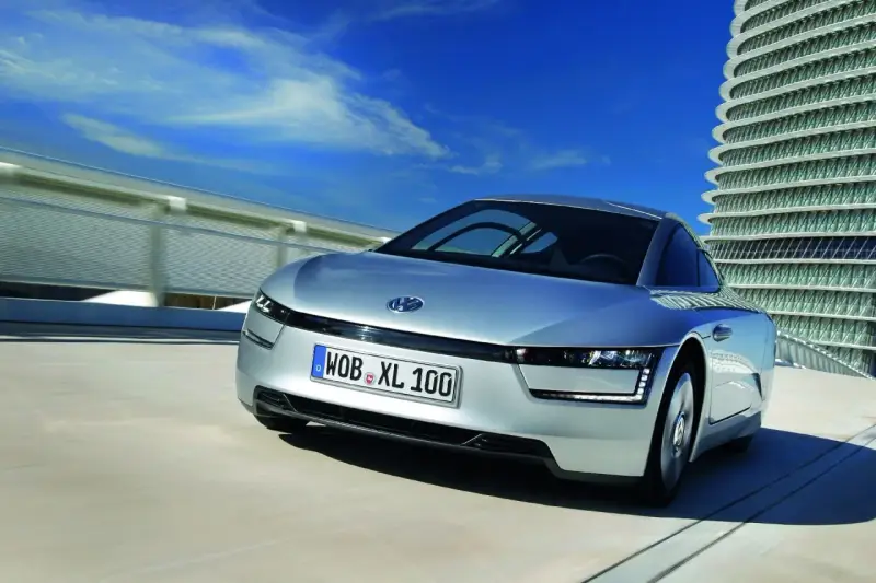 Srebrny, futurystyczny VW XL1 sunie po drodze obok nowoczesnego budynku.