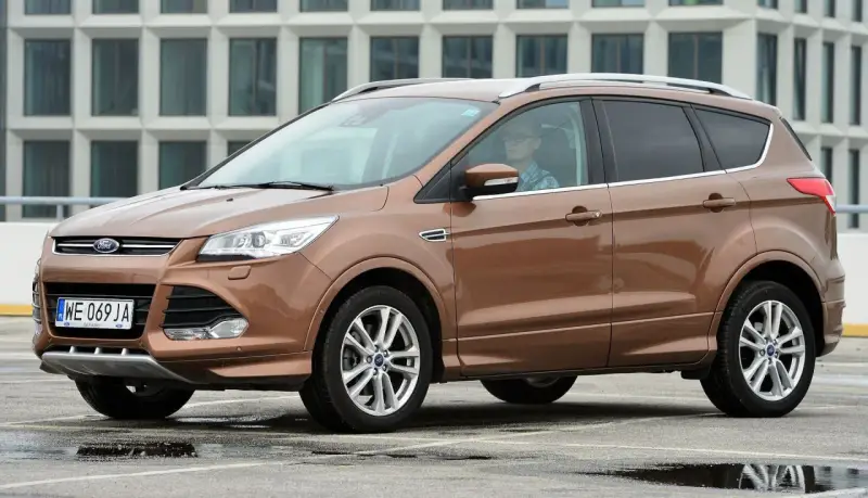 Brązowy Ford Kuga 2019 z kierowcą w okularach, zaparkowany na mokrym parkingu przed nowoczesnym budynkiem.