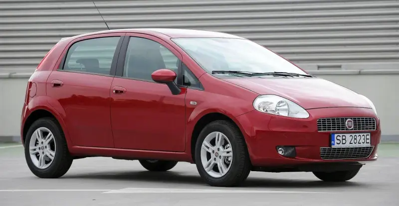 Czerwony Fiat Punto Evo z polskimi tablicami SB 2823E zaparkowany na szarym podłożu, z metalową ścianą w tle.